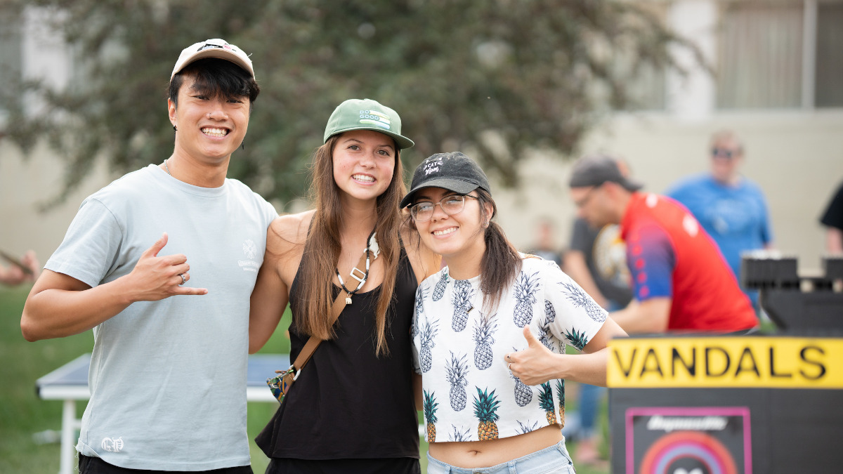 Smiling dark haired young man with baseball hat on head gives 'hang loose' hand gesture on grassy lawn with two smiling dark-haired young women in baseball caps.