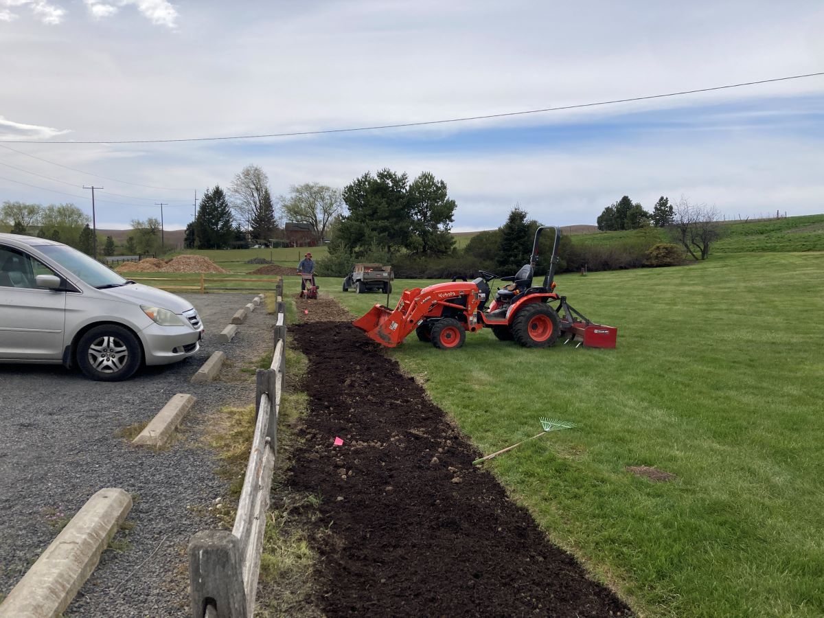 A tractor digging a flowerbed along the border of a parking lot.