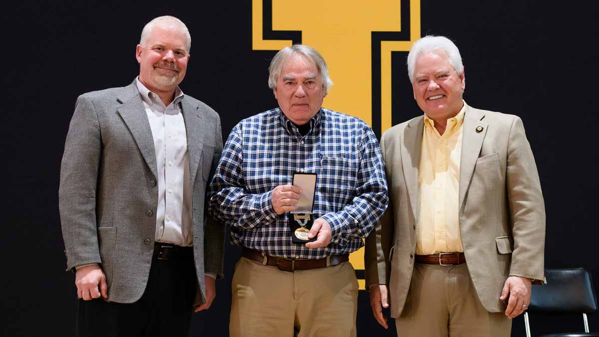 A group photo of three men, center man holding a medal.