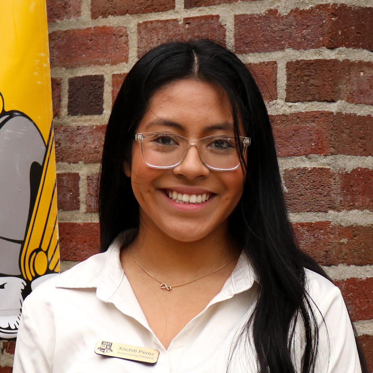 Xochitl Perez smiles outdoors on campus, a brick building in the background with a banner featuring an illustration of Joe Vandal.