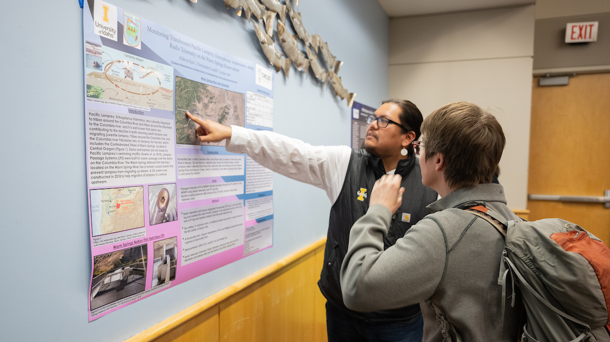 Man with chin beard and glasses points to a scientific poster while speaking to man wearing a backpack.