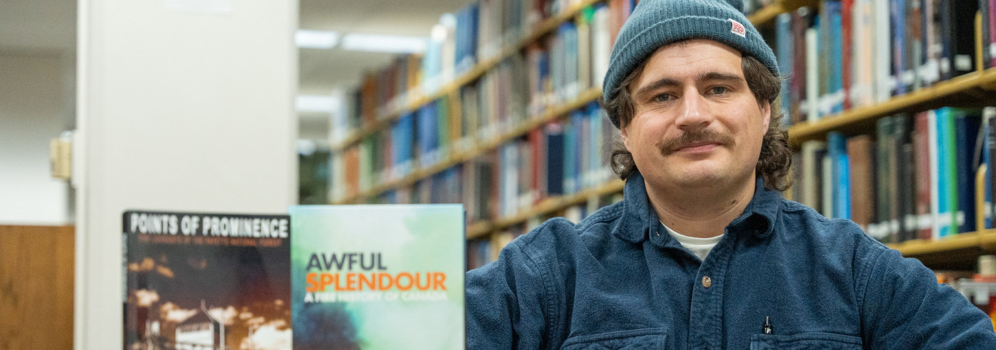 Man wearing woolen hat sits near two books displayed on table.