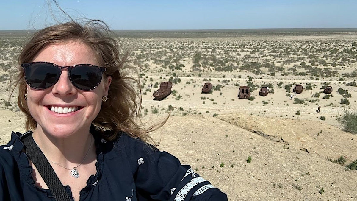 Woman wearing sunglasses with several rusty ships in a dry seabed behind her.
