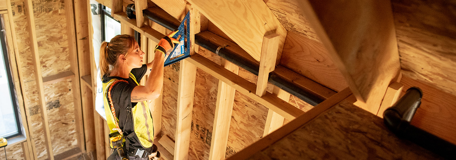 A woman measures something in the ceiling of a partially built wood house.