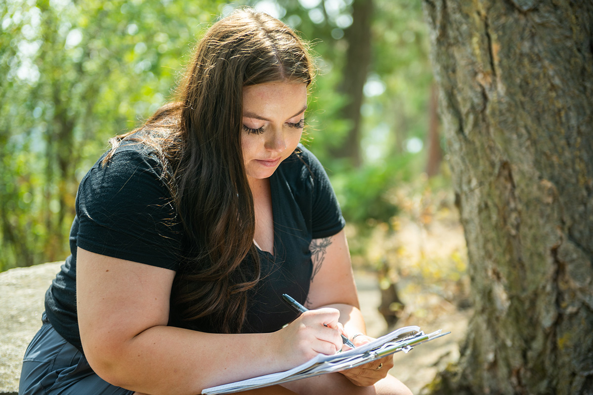 Emma Arman takes notes next to a tree in the forest.